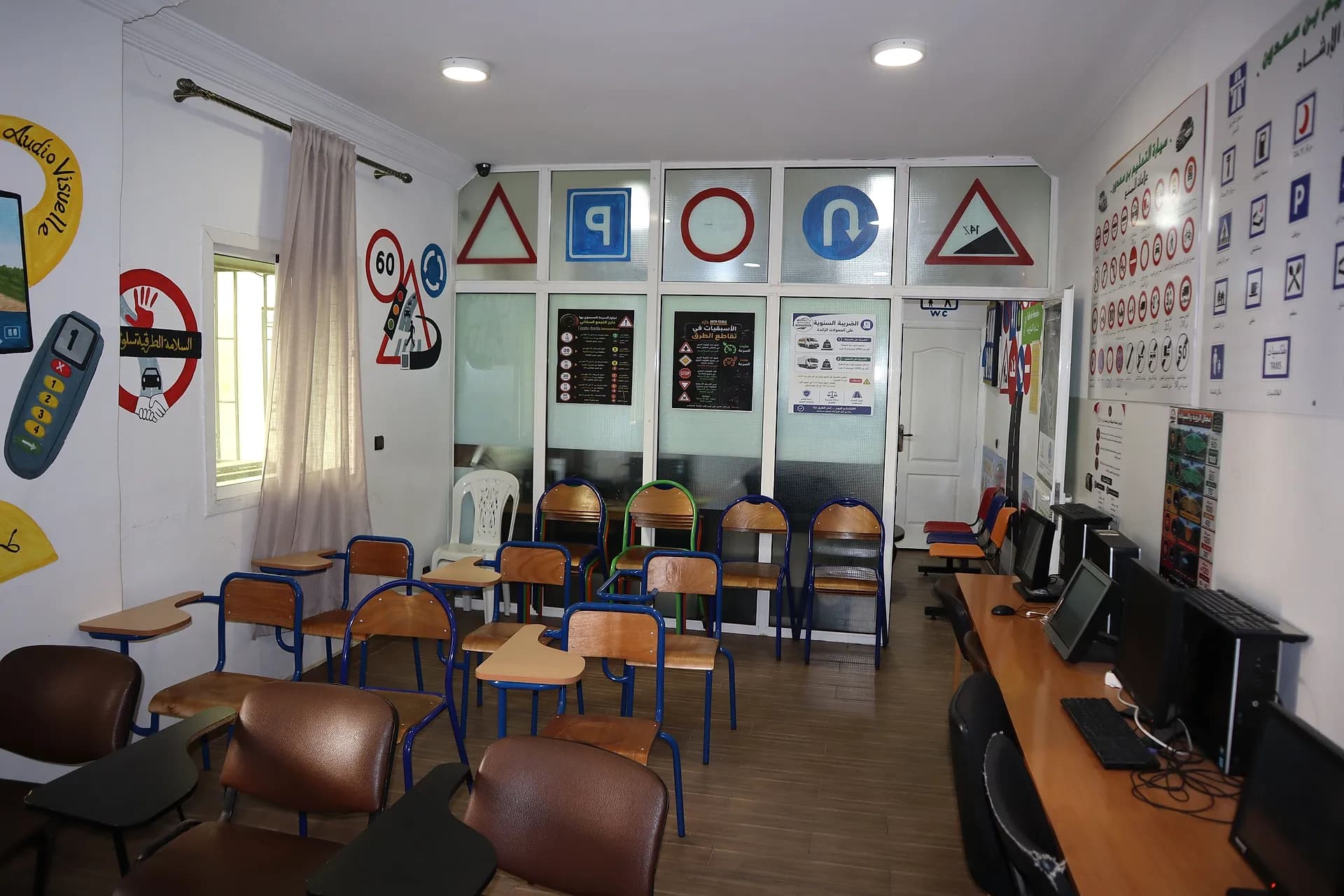 Salle de cours de l’auto-école face aux vitres décorées de panneaux, avec affiches du code de la route